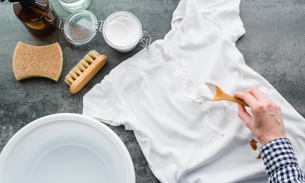 Eine Hand in kariertem Hemd streut Backpulver auf ein fleckiges weißes T-Shirt, umgeben von Bürste, Schwamm und Wasserbehälter. | © Adobe Stock/ HalynaRom
