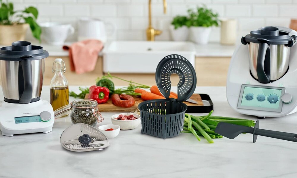Auf einer weißen Küchenarbeitsplatte stehen Thermomix-Geräte neben Zutaten wie Paprika, Karotten und einer Wurst auf einem Holzbrett. | © Vorwerk