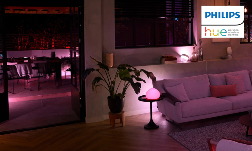 Wohnzimmer mit violettem Ambientelicht, grauem Sofa, Beistelltisch mit rosa Lampe und Blick auf beleuchtete Terrasse bei Nacht. | © Amazon/Philips