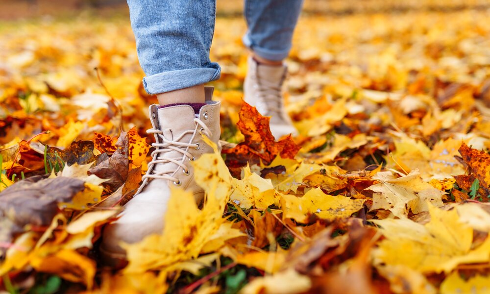 Beige Stiefel treten auf gelbe und braune Herbstblätter, die in einer Wiese verstreut liegen, dazu hochgekrempelte Jeans. | © Adobe Stock/ Olga