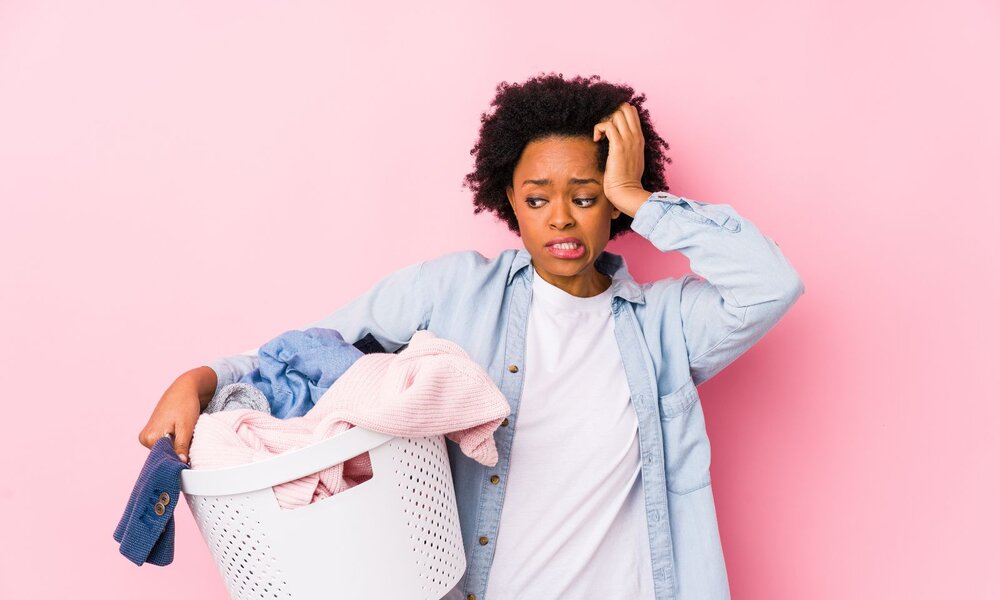 Junge Frau mit besorgtem Gesichtsausdruck hält einen vollen Wäschekorb vor rosa Hintergrund. | © Adobe Stock/ Asier