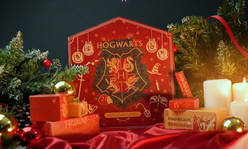 Roter Harry Potter-Adventskalender mit Hogwarts-Wappen, umgeben von Tannenzweigen, Kerzen und goldenen Weihnachtskugeln auf rotem Stoff. | © Amazon/Cinereplicas