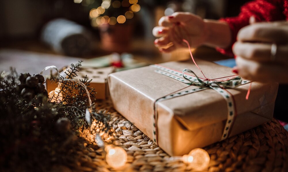 Weibliche Hände packen ein Geschenk mit Schleife vor einem Weihnachtsbaum ein, in Nahaufnahme. | © Getty Images / Pekic