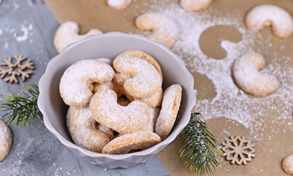 Mehrere Vanillekipferl in einer weißen Schüssel, daneben und darüber Puderzucker und weihnachtliche Deko. | © 	Firn