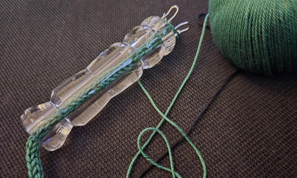 Eine durchsichtige Strickliesel mit grüner Wolle liegt auf einem dunklen, strukturierten Stoffhintergrund. | © Sabrina Forst