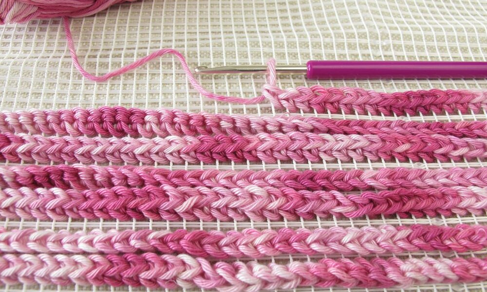 Nahaufnahme eines Stickgewebes mit rosa und pinken Garnreihen, daneben eine Häkelnadel mit pinkem Griff. | © Heike Rau / stock.adobe.com