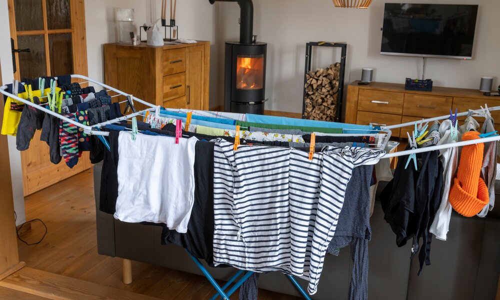 Ein Wäscheständer mit bunter Kleidung und Socken steht vor einem Kaminofen in einem gemütlichen Wohnzimmer mit Holzmöbeln. | © Adobe Stock/ gabort