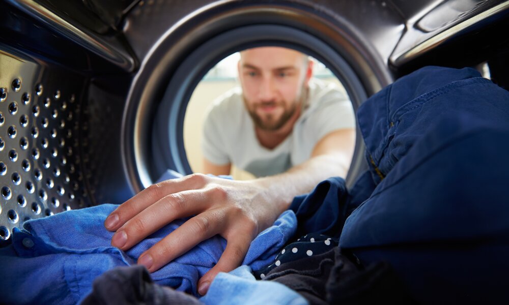 Ein junger Mann mit Bart greift konzentriert in eine Waschmaschine und sortiert blaue Kleidung vor hell beleuchtetem Hintergrund. | © Adobe Stock/ Monkey Business