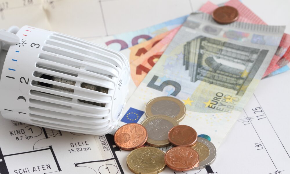 Ein weißer Heizungsregler liegt auf Bauplänen, umgeben von Euro-Scheinen und Münzen, symbolisch für Heizkosten und Energieplanung. | © Adobe Stock/ maho