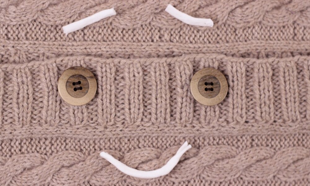Kreatives Gesicht aus Holzknöpfen und weißen Kordeln auf einem dicken, beigen Strickgewebe mit Zopfmuster. | © JuliaTim/stock.adobe.com