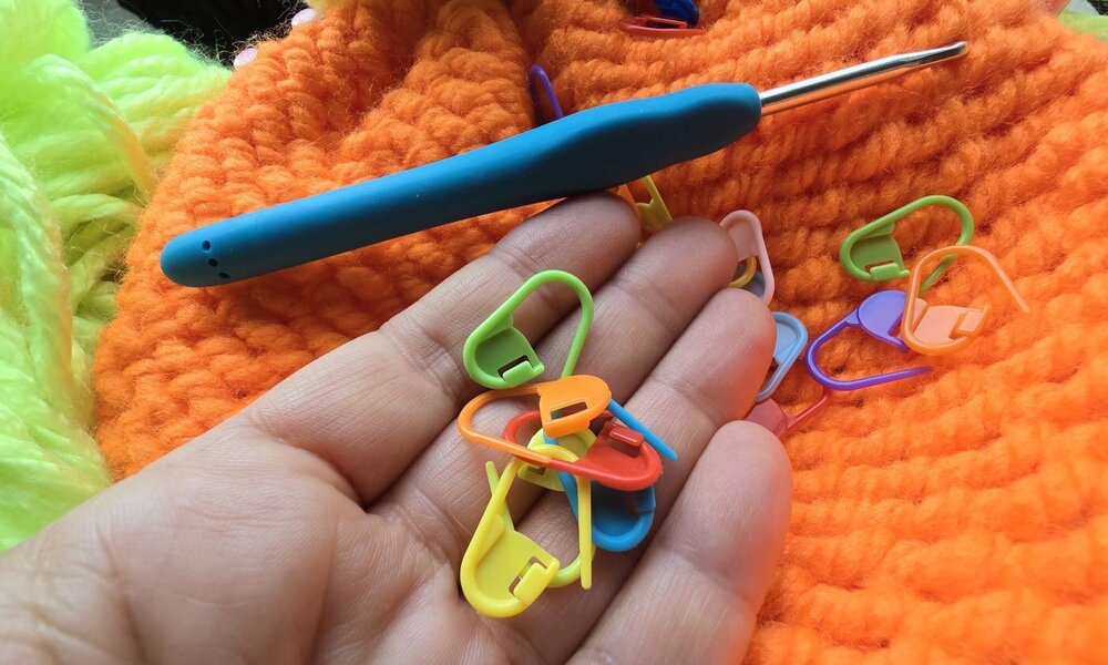 Eine Hand hält bunte Maschenmarkierer vor orangefarbener Wolle, daneben liegt eine blaue Häkelnadel auf der Strickarbeit. | © Sabrina Forst