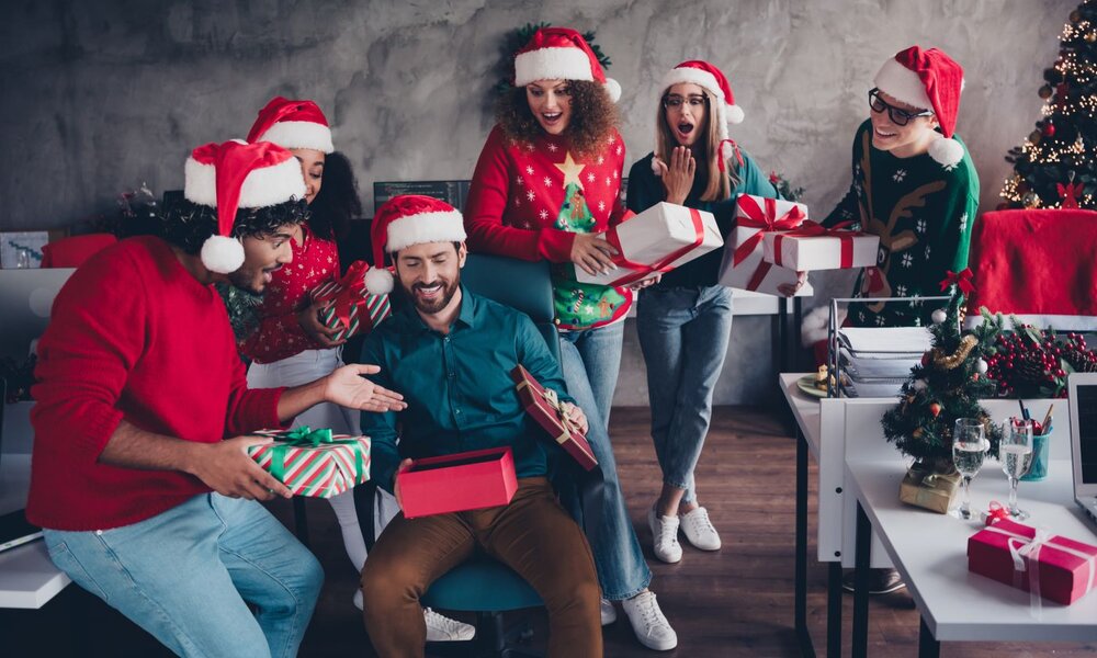 Eine fröhliche Gruppe junger Kollegen und Kolleginnen mit Weihnachtsmützen beschenkt sich lachend in einem festlich geschmückten Büro.  | © deagreez / stock.adobe.com