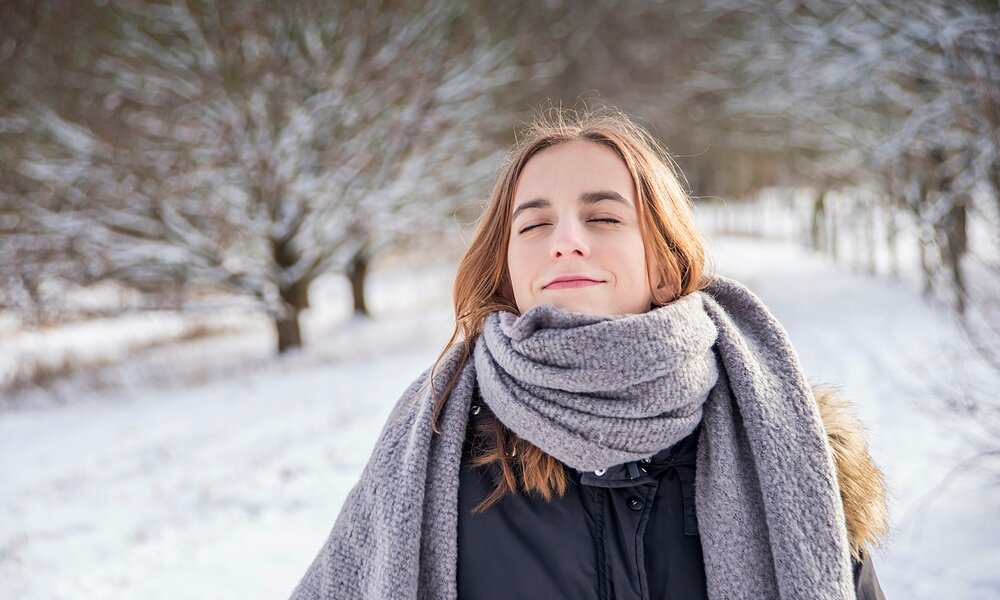 Junge Frau mit braunen Haaren und geschlossenen Augen genießt lächelnd einen sonnigen Wintertag in einem verschneiten Park. | © Adobe Stock / luna