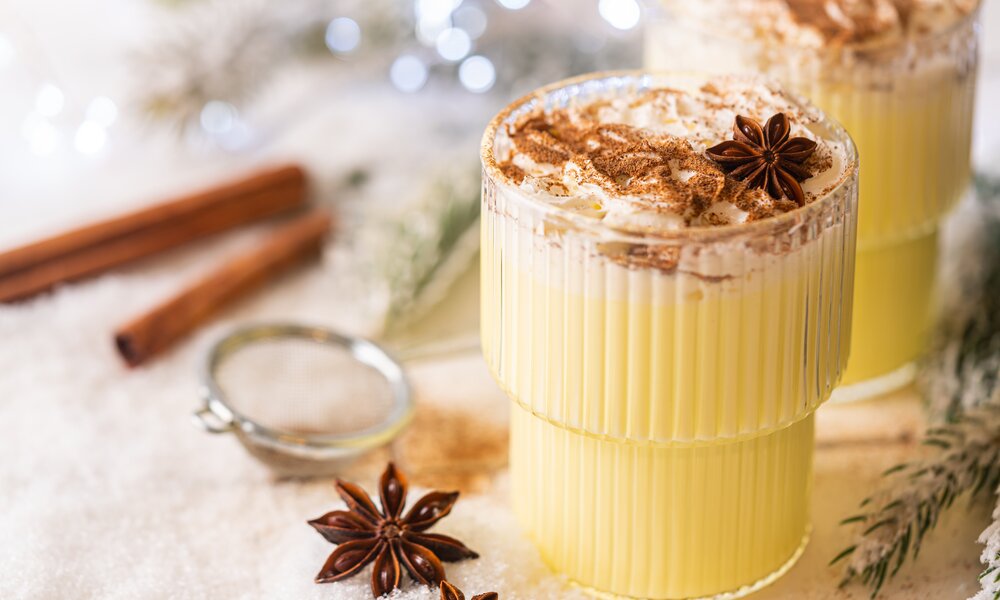 Zwei Gläser cremiger Eggnog mit Sahnehaube, Zimt und Sternanis stehen auf schneebedecktem Untergrund neben Zimtstangen. | © Adobe Stock/ petrrgoskov