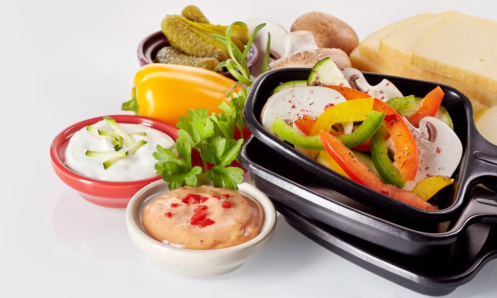 Frisches Gemüse wie Paprika, Zucchini und Champignons in Raclette-Pfännchen mit Dips, Gurken, Käse und Kräutern auf weißem Untergrund. | © Adobe Stock/ exclusive-design