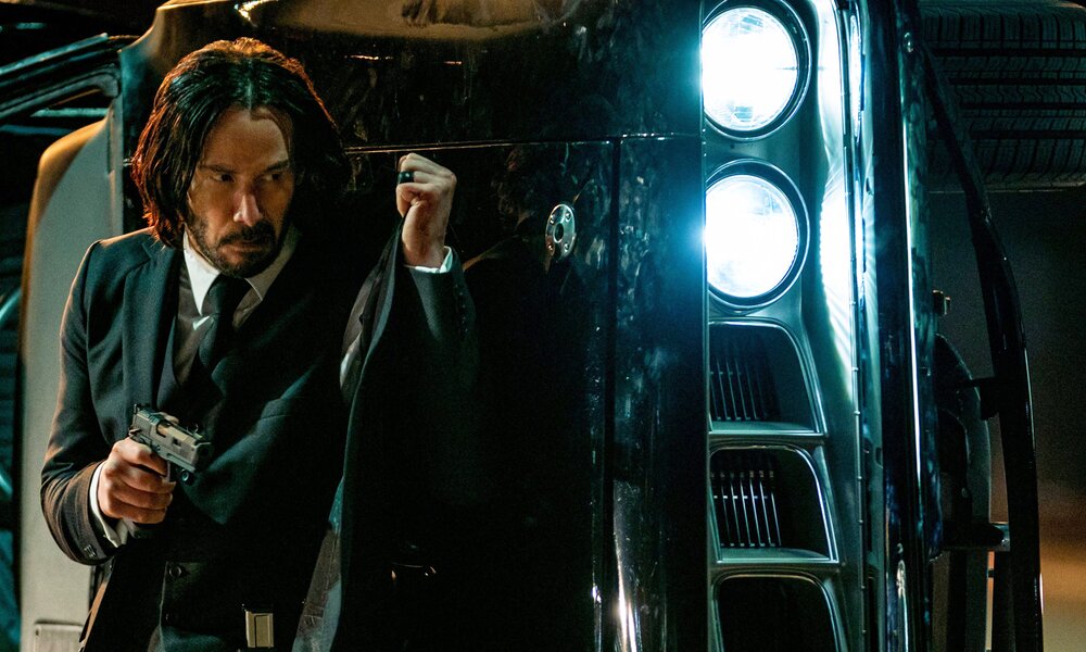Keanu Reeves als John Wick geht mit gezogener Waffe hinter einem Auto in Deckung in einer nächtlichen Szene aus dem Kinofiilm "John Wick: Kapitel 4" | © Imago Images/Murray Close/Capital Pictures