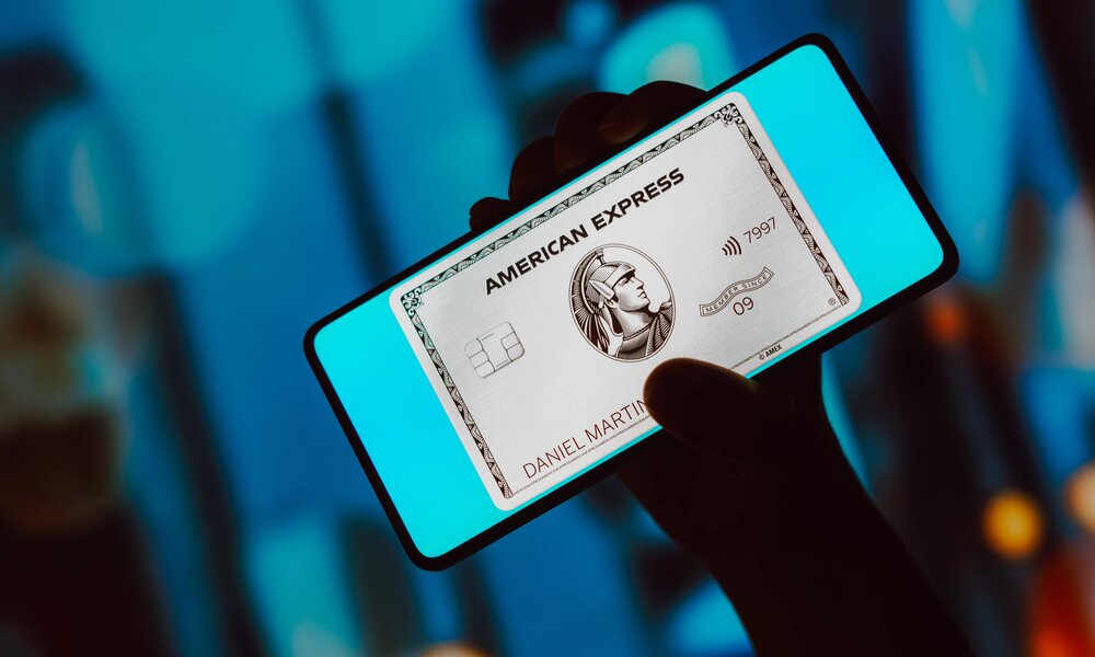 Smartphone mit leuchtendem Bildschirm zeigt silberfarbene American Express Kreditkarte, dahinter verschwommener Hintergrund in Blautönen. | © IMAGO/Zoonar