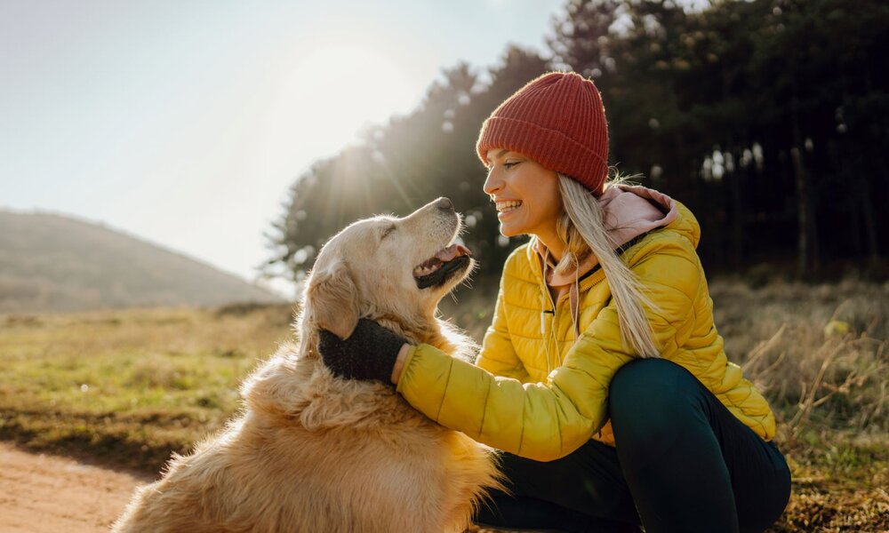 Eine junge Frau mit blonden Haaren und roter Mütze lächelt ihren Golden Retriever an, während sie ihn im Sonnenlicht streichelt. | © Getty Images / AleksandarNakic