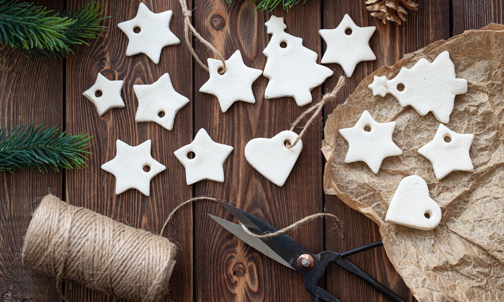 Selbstgemachte weiße Salzteig-Anhänger in Stern-, Herz- und Tannenbaumform liegen auf einer Holzfläche mit Zweigen und Juteschnur. | © Adobe Stock/ Елена Швецова