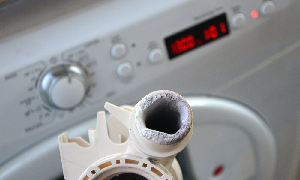 Ein stark verkalktes Waschmaschinen-Ablaufventil wird vor einer weißen Waschmaschine mit beleuchtetem Display und Drehknopf gehalten. | © Adobe Stock/ burnstuff2003