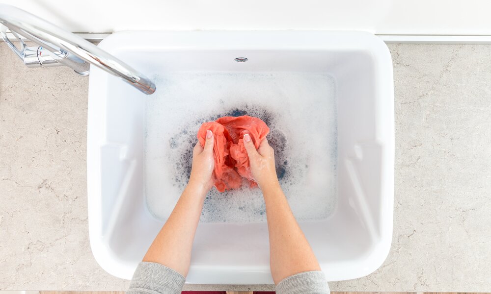 Eine Person mit hellen Händen wäscht ein rotes Kleidungsstück per Hand in einer weißen Spüle mit schäumendem Seifenwasser. | © Adobe Stock/ Mariakray