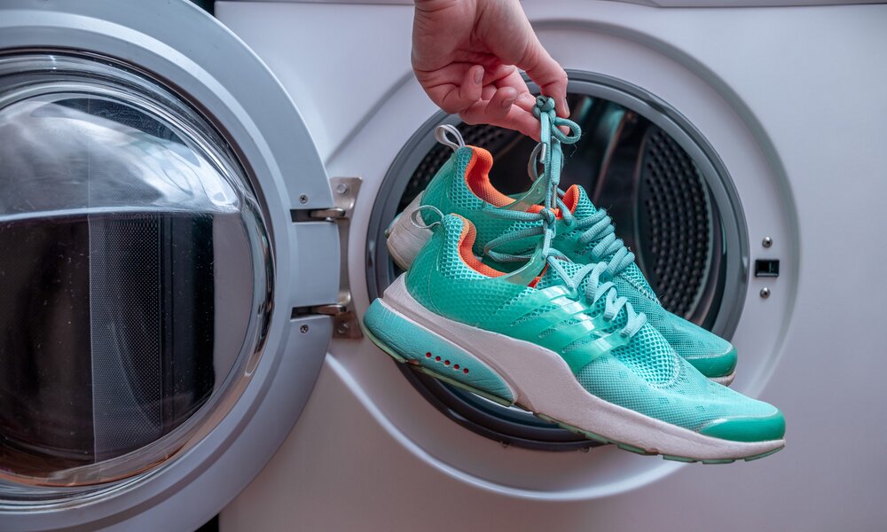 Eine Hand zieht türkisfarbene Sportschuhe mit orangefarbenem Innenfutter aus der geöffneten Trommel einer Waschmaschine. | © Adobe Stock/ Goffkein
