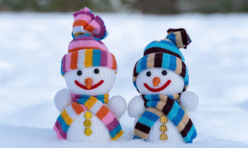 Zwei lächelnde Schneemänner mit bunter Strickmütze und Schal stehen nebeneinander auf schneebedecktem Boden vor verschwommenem Hintergrund. | © IhorStore / stock.adobe.com
