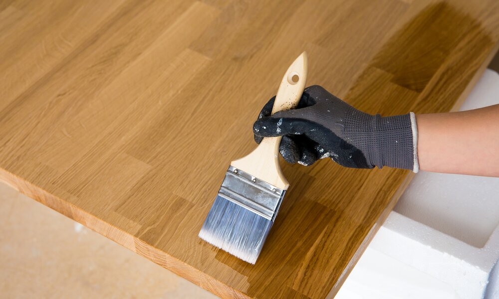 Nahaufnahme eines lackierten Pinsels, gehalten von einer Hand in Handschuhen, beim Versiegeln einer Holzoberfläche. | © Adobe Stock/ FotoHelin