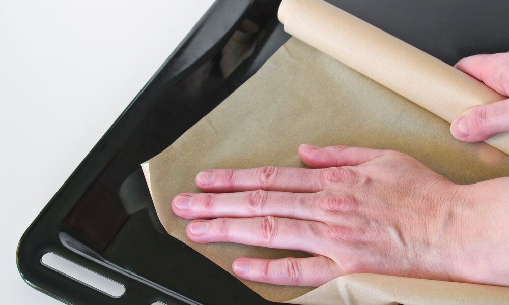 Backpapier auf schwarzem Backblech bildfüllend, Hand streicht Backpapier glatt auf das Blech | © Adobe Stock/Gehkah