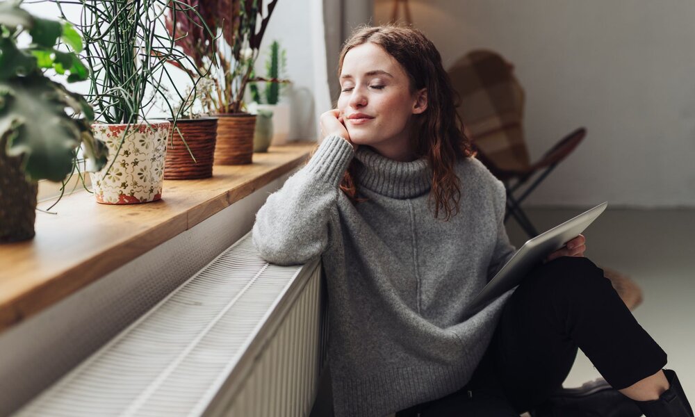 Frau mit geschlossenen Augen und grauem Rollkragenpullover sitzt lächelnd an einer Heizung neben einer Fensterbank mit Pflanzen. | © Adobe Stock/ contrastwerkstatt