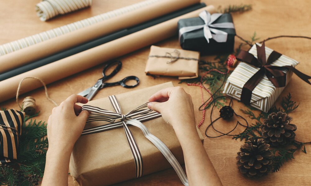 Hände einer Person binden eine gestreifte Schleife um ein in braunes Papier eingepacktes Geschenk auf einem Tisch mit Tannenzweigen und Kiefernzapfen. | © Adobe Stock/ sonyachny