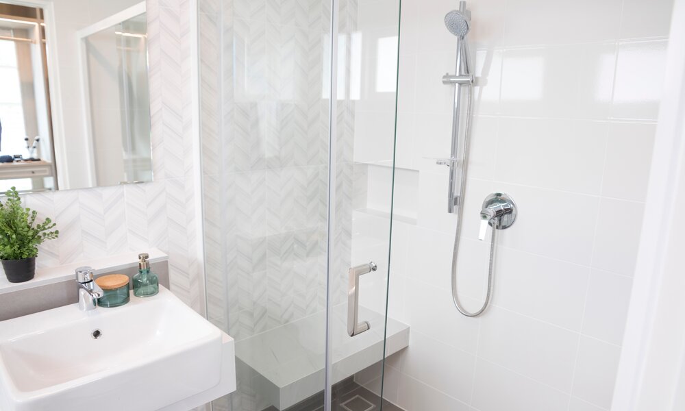 Helles Badezimmer mit moderner Glasdusche, weißer Wandverkleidung, kleiner Duschablage und einem Waschbecken mit Deko. | © Adobe Stock/ bennnn