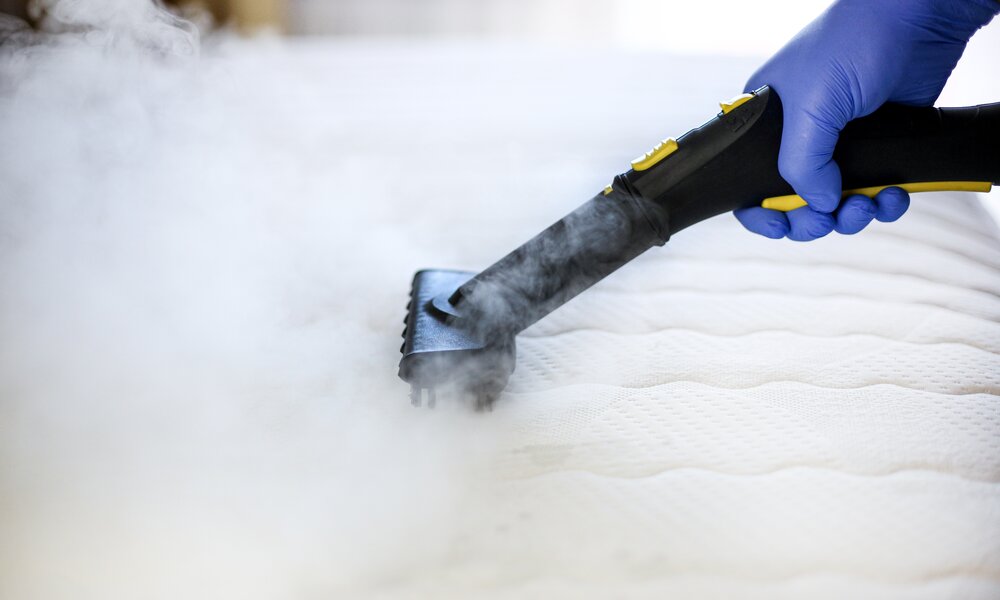 Eine Hand in blauen Einweghandschuhen reinigt mit einem Dampfreiniger eine weiße, strukturierte Matratze, aus der Dampf aufsteigt. | © Adobe Stock/ Denis Barysau