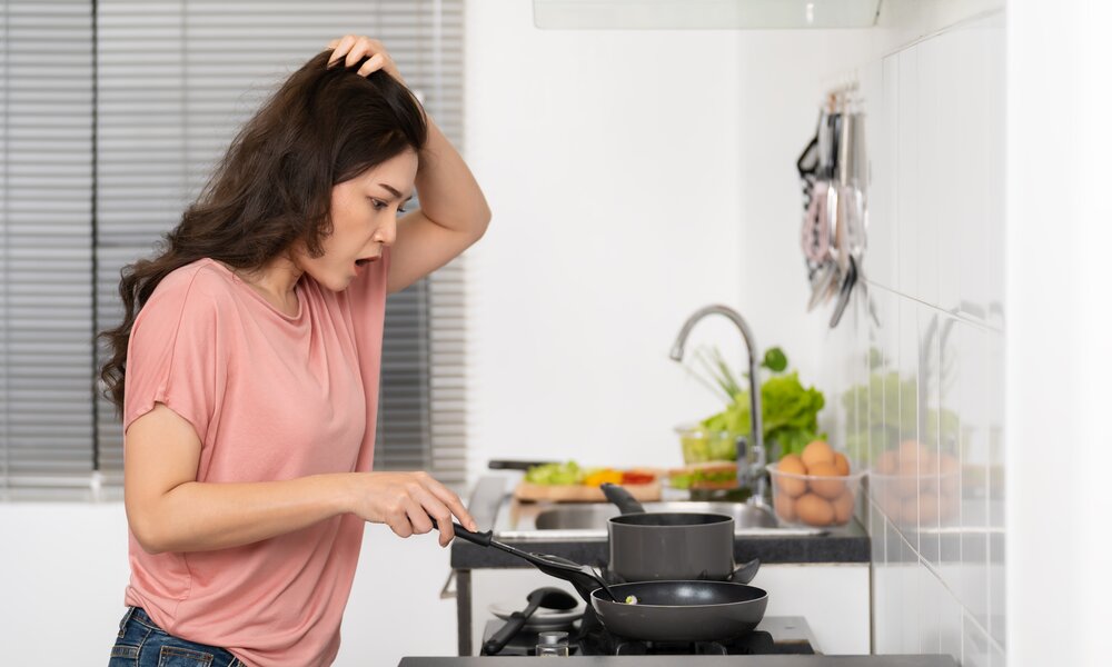 Verwirrte Frau in Jeans und rosa Oberteil greift sich an den Kopf, während sie in einer Pfanne in einer hellen Küche kocht. | © Adobe Stock/ geargodz