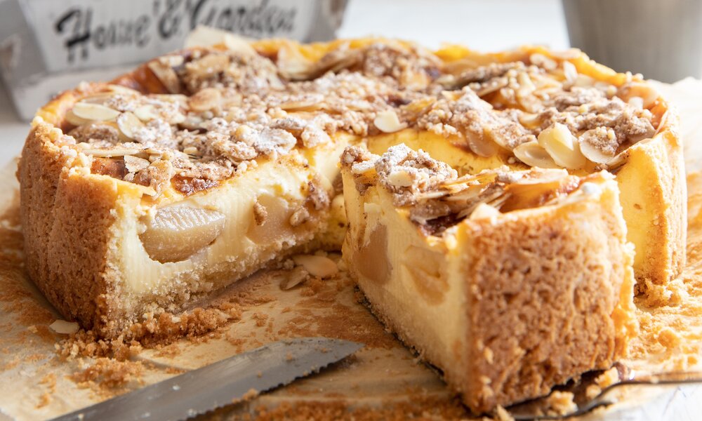 Angeschnittener Apfel-Schmand-Kuchen auf einem Holztisch, daneben frische Äpfel. | © Adobe Stock / Kerstin