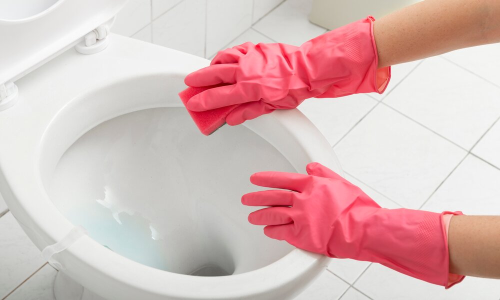 Zwei Hände in pinken Gummihandschuhen reinigen mit einem roten Schwamm den weißen Toilettenrand in einem gefliesten Badezimmer. | © Adobe Stock/ Impact Photography
