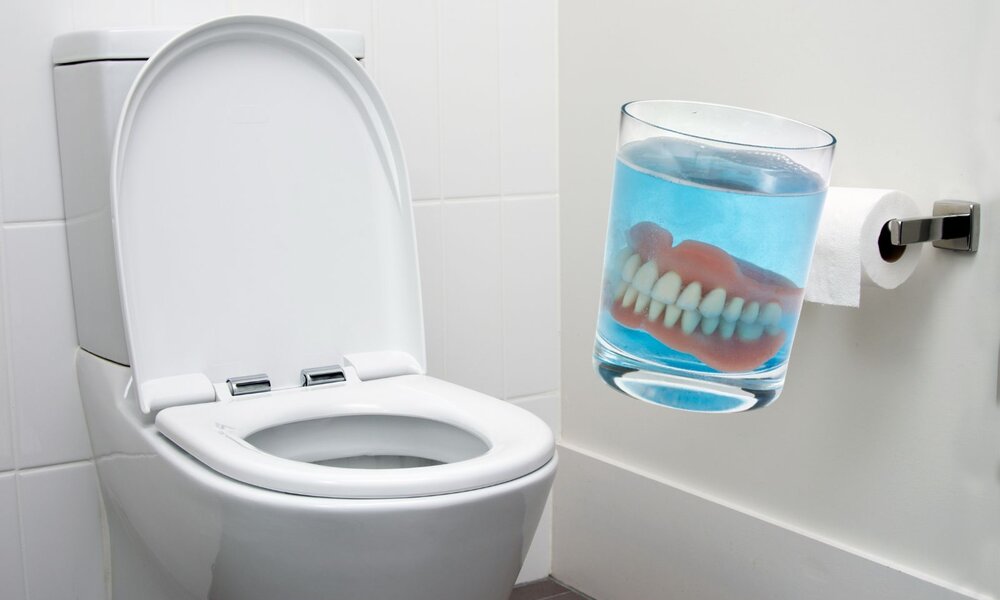 Neben einer weißen Toilette und einer Toilettenpapierrolle steht ein Glas mit einer Zahnprothese in blauer Reinigungsflüssigkeit. | © Adobe Stock/ gmstockstudio/ fotofuerst