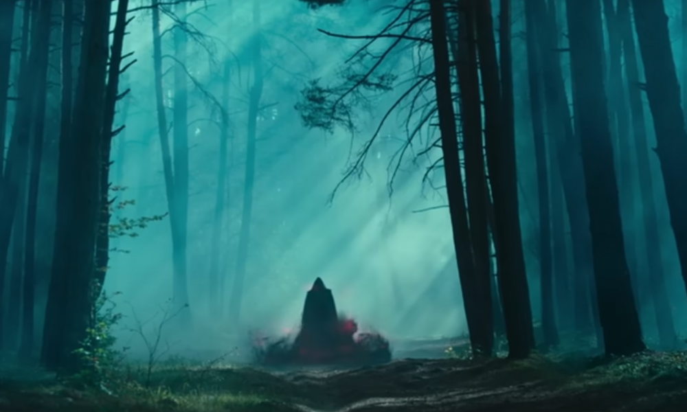 Eine dunkle, mystische Gestalt mit Kapuze steht in einem nebligen Wald, umgeben von roten Rauchwolken und Sonnenstrahlen. | © Sony Pictures Entertainment / Screenshot Trailer "Dampyr"
