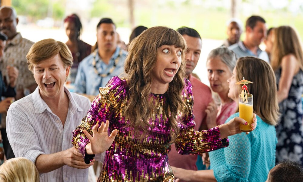 Eine junge Frau mit langem braunem Haar in glitzerndem pink-goldenem Kleid macht einen erschrockenen Gesichtsausdruck, während sie einen Cocktail hält. | © Katrina Marcinowski/NETFLIX