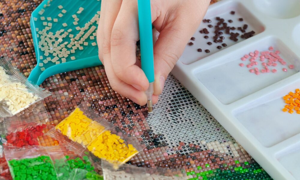 Eine Hand platziert mit einem türkisfarbenen Stift winzige Mosaiksteinchen auf eine Vorlage, umgeben von bunten Steinchen in Tüten. | © Skiminok/ stock.adobe.com