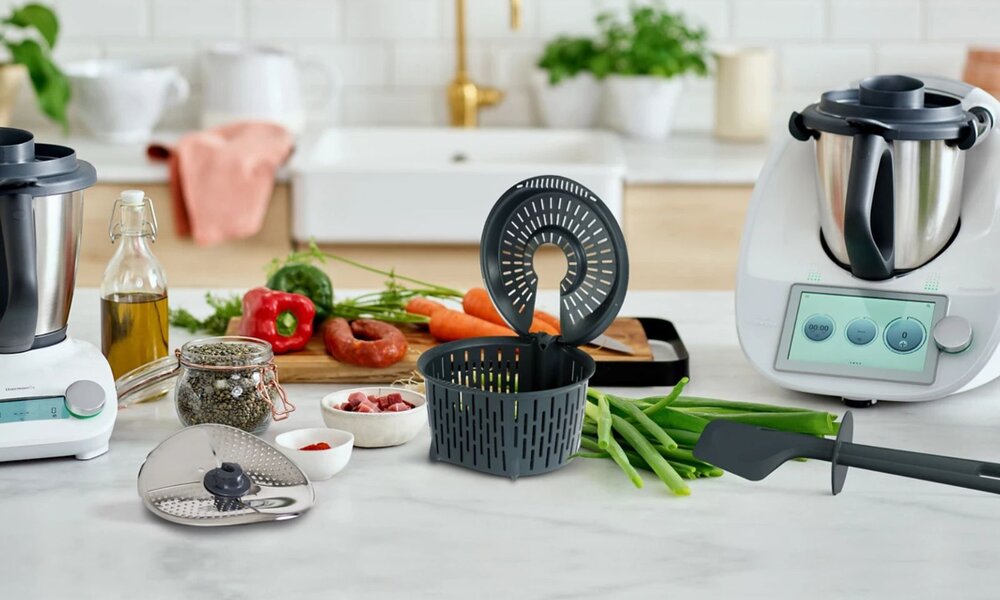 Thermomix-Gerät mit Zubehör steht auf einer Arbeitsplatte, umgeben von frischen Zutaten wie Gemüse und Gewürzen. | © Vorwerk