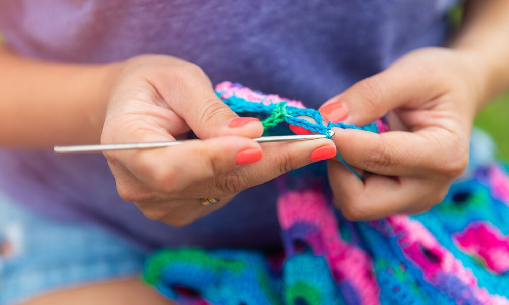 Eine Frau in lila Shirt häkelt ein buntes Tuch in kräftigen Farben, die Hände präzise und konzentriert im Fokus. | © manuta/ stock.adobe.com