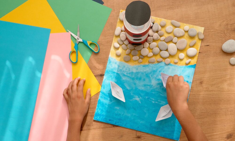 Ein Kind bastelt auf einer Holztischplatte eine Strandlandschaft aus Papier, Steinen und Origami-Booten, neben buntem Bastelpapier und einer Schere. | © vejaa/ stock.adobe.com