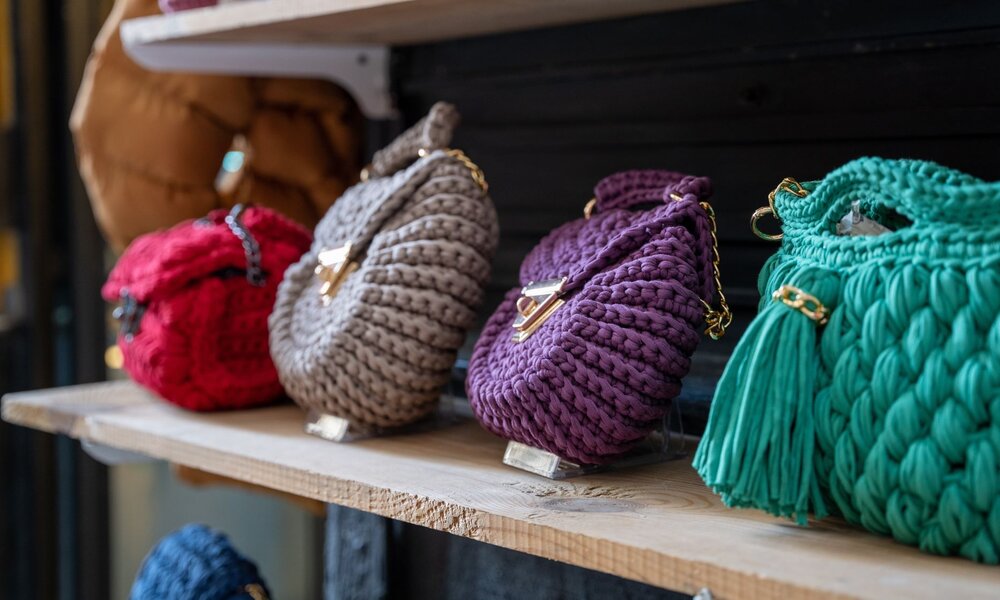 Vier handgefertigte Häkelhandtaschen in Rot, Beige, Lila und Grün mit goldenen Kettenhenkeln stehen auf einem Holzregal. | © Erman Gunes / stock.adobe.com