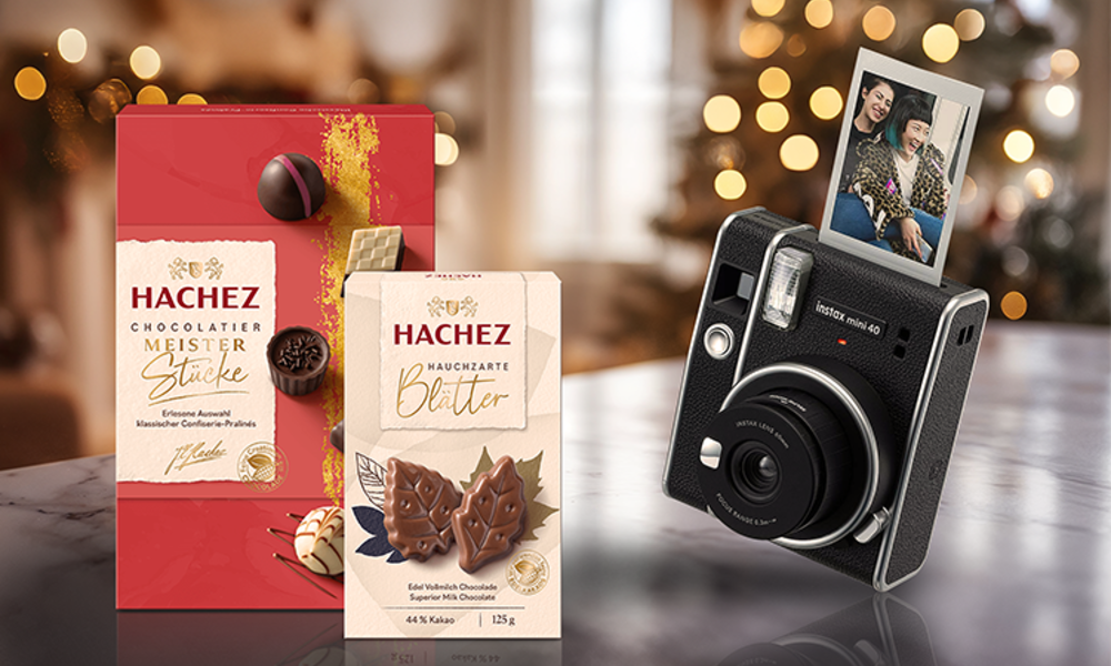 Hachez-Schokolade in roten und beigen Verpackungen mit Pralinen und Blättern, daneben eine Kamera mit Polaroid vor festlicher Dekoration. | © Hachez