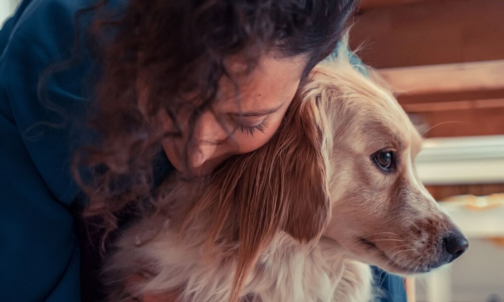 Eine junge Frau mit dunklen Locken umarmt zärtlich einen cremefarbenen Hund mit sanftem Blick in einem gemütlichen Innenraum. | © Getty Images / erondell