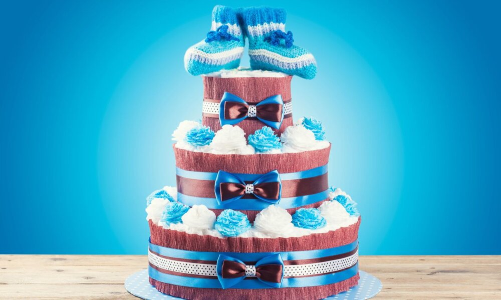 Dreistöckige Windeltorte mit blauen Schleifen und Stoffblumen, oben dekoriert mit gestrickten Babyschuhen auf blauem Hintergrund. | © merydolla/ stock.adobe.com