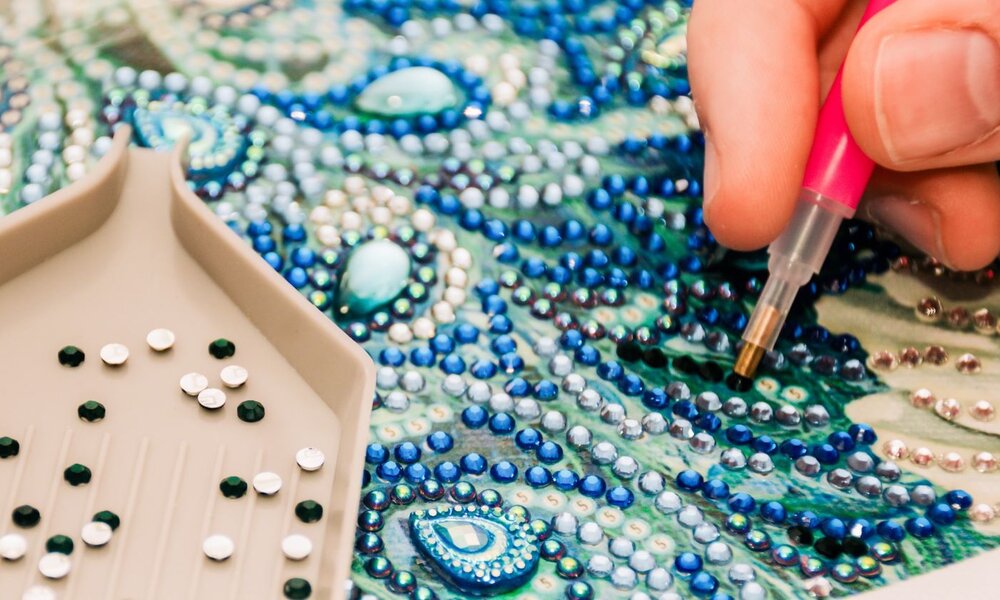 Eine beige Schale mit glitzernden Strasssteinen neben einer Hand, die ein detailreiches Kunstwerk aus Schmucksteinen gestaltet. | © iStock / Getty Images Plus / Daria Nipot