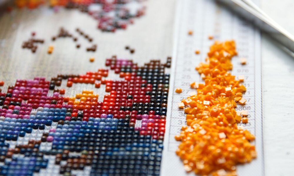 Nahaufnahme eines halbfertigen Diamond-Painting-Motivs mit farbigen Steinen, gelben Perlen und Pinzette auf einem Raster. | © Mariana Rusanovschi /stock.adobe.com