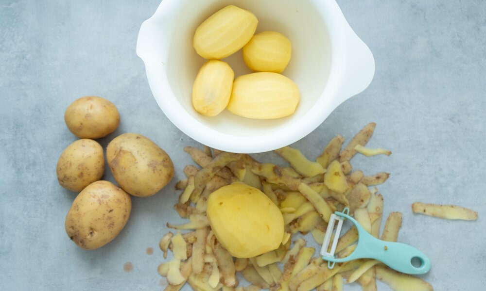 Geschälte Kartoffeln in einer weißen Schüssel, daneben ungeschälte Kartoffeln, Schalen und ein türkiser Sparschäler auf grauem Untergrund. | © Adobe Stock/ Anastasia
